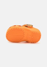 Orange gummisål med et tekstureret mønster. Har perforerede sider og en rem med en justerbar spænde.