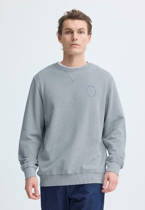 Felpa grigia con collo e polsini a costine, caratterizzata da un piccolo disegno circolare sul petto, indossata sopra una canottiera bianca.