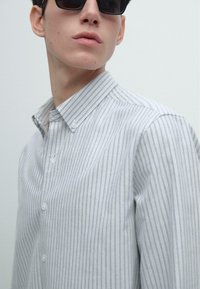 Koszula zapinana na guziki w jasnoszarym kolorze z pionowymi białymi paskami, posiadająca standardowy kołnierzyk i teksturowaną bawełnianą tkaninę. Subtelny detal w postaci guzików.