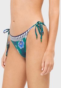 Femme portant un bas de bikini imprimé floral vert, bleu et blanc avec des liens latéraux, montrant la taille et le haut des cuisses.
