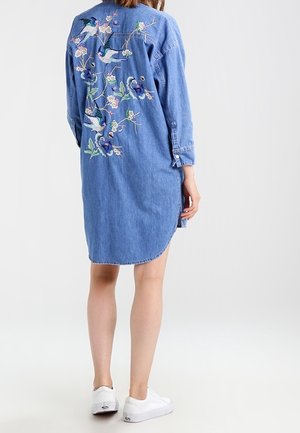 Farkkupaitamekko, jossa on pitkät hihat ja värikkäitä kirjailtuja lintuja ja kukkia selässä, yhdistetty valkoisten tennarien kanssa.