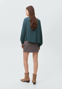 Blusa verde, ampia e comoda, con maniche lunghe, abbinata a una gonna di jeans grigia. Stivali alla caviglia marroni completano l'outfit. Sfondo neutro.
