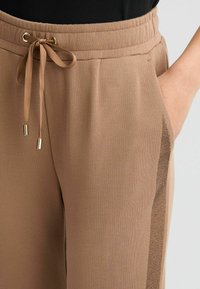 Braune Sweatpants mit einem Kordelzugbund und goldfarbenen Akzenten. Sie verfügen über ein seitliches Netzpaneel für zusätzlichen Textur- und Luftdurchlässigkeit.