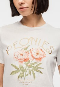 Femme portant un t-shirt blanc avec des fleurs de pivoine roses imprimées et le texte doré "PEONIES" au-dessus des fleurs à l'avant.