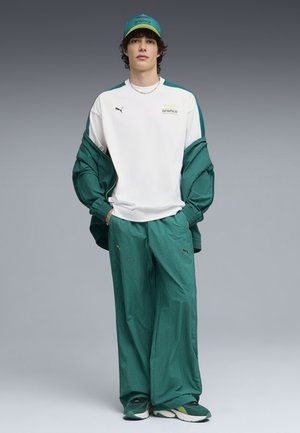 Jongere draagt een groen Puma trainingspak, een witte Aramco trui, een bijpassende groene pet en sneakers, staand met handen in de zakken tegen een grijze achtergrond.