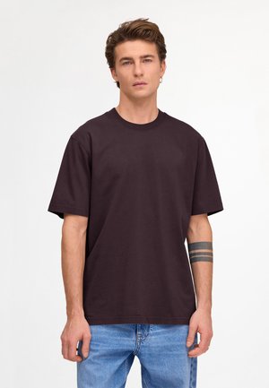 Ung mand med kort brunt hår iført en almindelig sort T-shirt og lyseblå jeans, der viser en sort tatovering med tre bånd på sin venstre underarm.