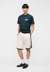 Ανδρική πράσινη και ναυτικού μπλε ριγέ μπλούζα με στρογγυλή λαιμόκοψη, σε συνδυασμό με κρεμ σορτς με σκουροπράσινες λεπτομέρειες και λογότυπο. Μαύρα παπούτσια και λευκές κάλτσες.