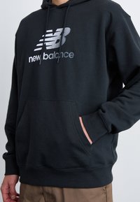 Črna kapuca iz mehkega materiala, s sprednjo žepno, rebrastimi manšeti in velikim odsevnimi logotipom "New Balance" na prsih.