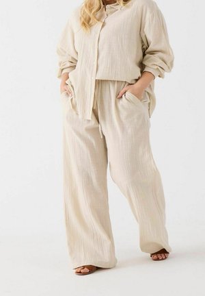 Kobieta ubrana w luźną beżową lnianą koszulę i spodnie o szerokich nogawkach, z rękami w kieszeniach, stoi na białym tle w brązowych sandałach.