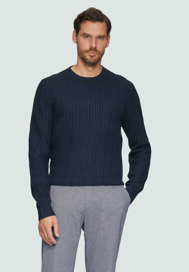 Granatowy sweter z dzianiny w sploty warkoczowe, z okrągłym dekoltem i długimi rękawami, z teksturowanym wzorem i ściągaczami przy mankietach, w połączeniu z szarymi spodniami.