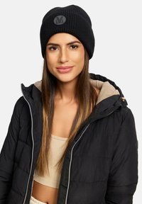 Berretto in maglia nera con texture a coste e un'applicazione circolare nera con logo sulla parte anteriore. Indossato con una giacca nera con zip e un top beige.