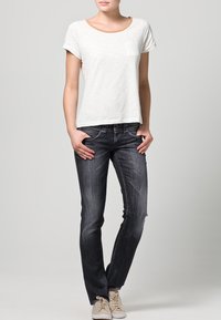 Camiseta blanca de manga corta con un bolsillo, combinada con jeans ajustados de lavado oscuro y zapatillas ligeras. El fondo es de un gris neutro.