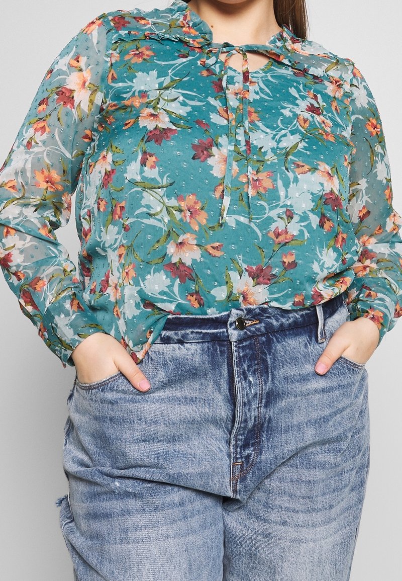 Blouse florale transparente turquoise avec col froncé et détail à nouer, ornée d'un mélange de fleurs roses et blanches. Portée avec des jeans déchirés.