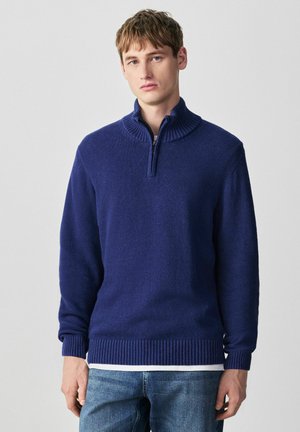 Young man wearing a blue quarter-zip sweater over a white shirt and blue jeans, standing against a plain light gray background.