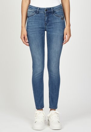 Schlanke blaue Jeans aus Denim, getragen mit weißen Turnschuhen und einem weißen Oberteil, gezeigt von der Mitte des Oberkörpers bis zu den Füßen vor einem einfarbigen Hintergrund.