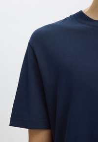 Camiseta azul marino de manga corta con cuello redondo, llevada por una persona visible desde el hombro hasta la mitad del pecho sobre un fondo liso.