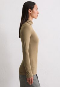 Frau mit langen dunklen Haaren, die einen engen beigen Rollkragenpullover und einen grauen Rock trägt, im Profil vor einer weißen, strukturierten Wand stehend.