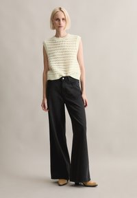 Strikket ermeløs topp i lys gul med et åpent mønster, kombinert med høy midje, vide svarte jeans og beige hæler.