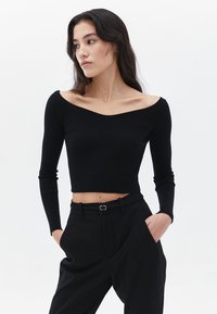 Schwarzes, geripptes Off-Shoulder-Oberteil mit langen Ärmeln, verkürztem Design, enganliegendem Schnitt und strukturierter Oberfläche. Kombiniert mit hochgeschnittenen schwarzen Hosen.