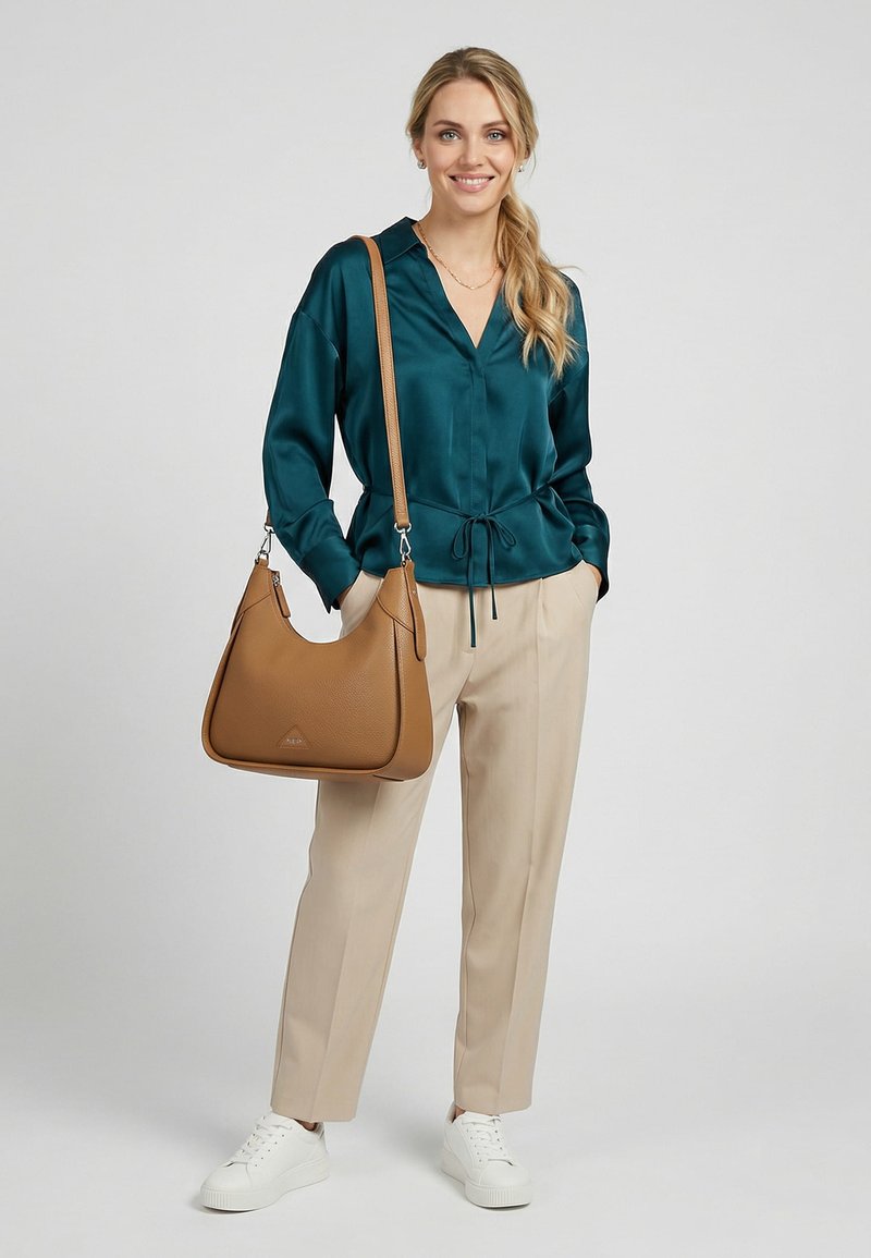 Mujer sonriente con cabello rubio, blusa verde azulado y pantalones beige, lleva un bolso grande marrón claro al hombro y usa zapatillas blancas, de pie sobre un fondo gris.