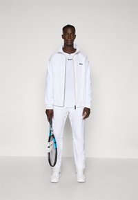 White athletic tracksuit featuring a zip-up jacket with a logo on the chest, paired with matching pants. Wielding a tennis racket.