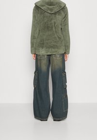 Person som bär en grön fluffig jacka och löst sittande blå denim cargobyxor med flera fickor, står vänd bort mot en enfärgad bakgrund.