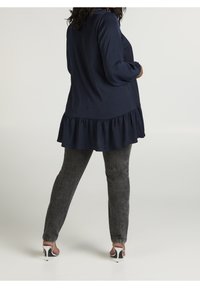 Mörkblå långärmad topp med rynkad nederkant, i kombination med grå texturerade byxor och silverfärgade högklackade skor. Enkel, lager-på-lager design.