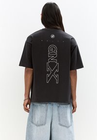 Black cotton t-shirt with a round neckline and short sleeves. Features large white "GNX" text vertically on the back, above a circular logo.