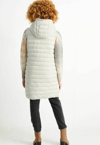 Osoba z kręconymi włosami, nosząca jasnoszary ocieplany kamizelka z kapturem, wielokolorowy sweter, ciemne spodnie i beżowe botki za kostkę, zwrócona plecami.