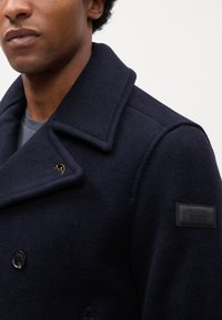 Marineblauer Wollmix-Peacoat mit doppelter Knopfleiste, breiten Revers und einem gebrandeten Lederpatch am Ärmel. Goldene Akzentdetails sind vorhanden.