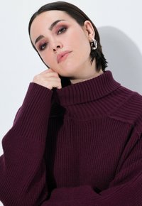 Burgundowy sweter z golfem, wykonany na dziewiarskiej maszynie, o prążkowanej fakturze i oversize'owym kroju. Posiada wysoki kołnierz oraz obniżone szwy na ramionach.