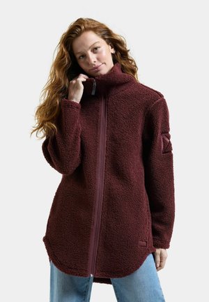 Vrouw met lang golvend haar, gekleed in een bordeauxrode fleecejas met rits en blauwe spijkerbroek, houdt met één hand de kraag van de jas vast.