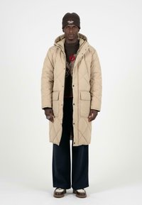 béžový prošívaný dlouhý kabát s kapucí, se zapínáním na patentky, dvěma předními kapsami, nošený přes tmavý svetr a kalhoty širokého střihu.