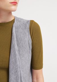 Chaleco gris sin mangas sobre una camiseta de punto acanalado mostaza, que muestra una textura suave con un diseño drapeado y mangas ajustadas.