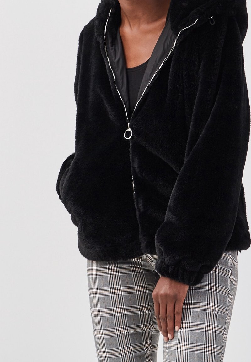 Personne portant une veste noire duveteuse à fermeture éclair avec des poignets élastiques et un pantalon à carreaux gris, debout, une main détendue le long du corps.