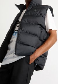 Svart pufferväst med dragkedja, hög krage och logotyp. Bärs över en grå T-shirt och visar en quiltad design och slät textur.