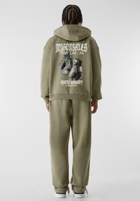 Olivgrüner Hoodie mit einem großen Grafikdruck von Boxhandschuhen auf dem Rücken, versehen mit weißem Text darüber und darunter. Kombiniert mit passender Jogginghose.