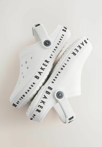 Białe chodaki z perforowanym wzorem, wyposażone w czarną opaskę z logo wzdłuż podeszwy oraz regulowany tylny pasek. Gładkie i teksturowane powierzchnie.