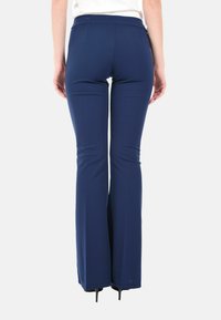 Donna che indossa pantaloni a zampa di elefante blu navy e tacchi alti neri, vista di spalle su uno sfondo bianco.