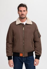 Hombre con cabello corto y castaño y bigote lleva chaqueta bomber marrón con cuello beige y vaqueros azules, de pie frente a un fondo blanco liso.