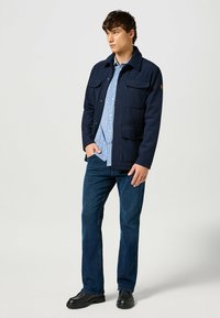 Chaqueta de mezcla de lana en color navy con dos bolsillos frontales, cierre de botones y cuello, llevada sobre una camisa a cuadros en color azul claro, combinada con jeans oscuros y botas negras.