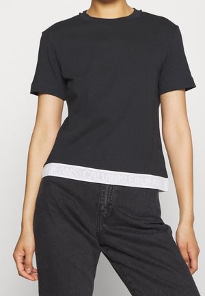 Person wearing a black short-sleeve T-shirt with a white Calvin Klein Jeans logo band at the hem, paired with black jeans.