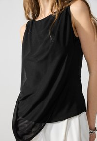 Black sleeveless top made of smooth fabric, featuring a draped design with an asymmetrical hem that subtly layers over a white garment.
