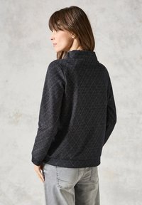 Sort mønstret jakke med en diamantdesign, høj krave, lange ærmer og en afslappet pasform. Båret med lysegrå jeans.