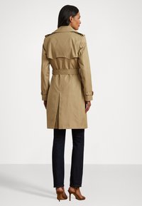 Beige trench coat with shoulder epaulets and a belted waist, featuring a back vent and long sleeves, paired with dark jeans and brown heels.