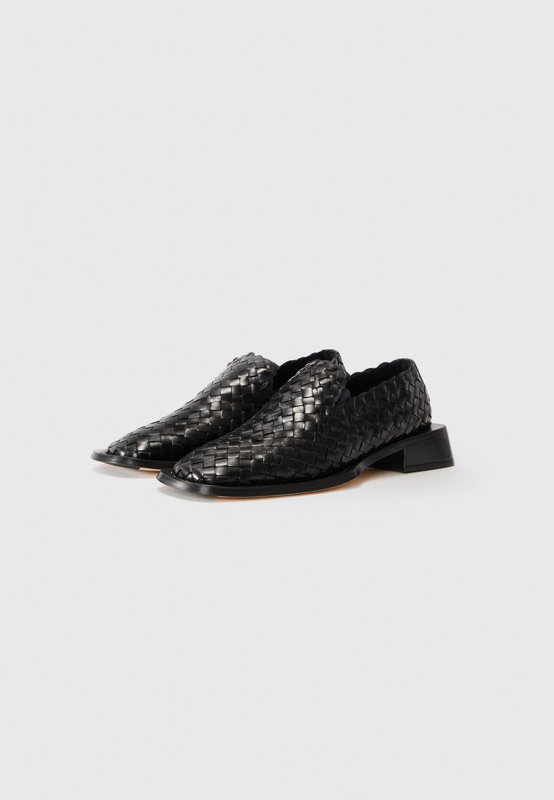 Een paar zwarte leren gevlochten instap-schoenen met lage blokhakken op een effen witte achtergrond.