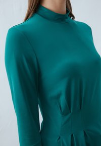 Teal kjole med høy krage, lange ermer og plisserte detaljer ved livet i glatt, lett stoff.