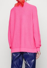 Ljust rosa stickad tröja med ett texturerat mönster, ribbade ärmslut och en lös passform, i kombination med glänsande marinblå byxor.