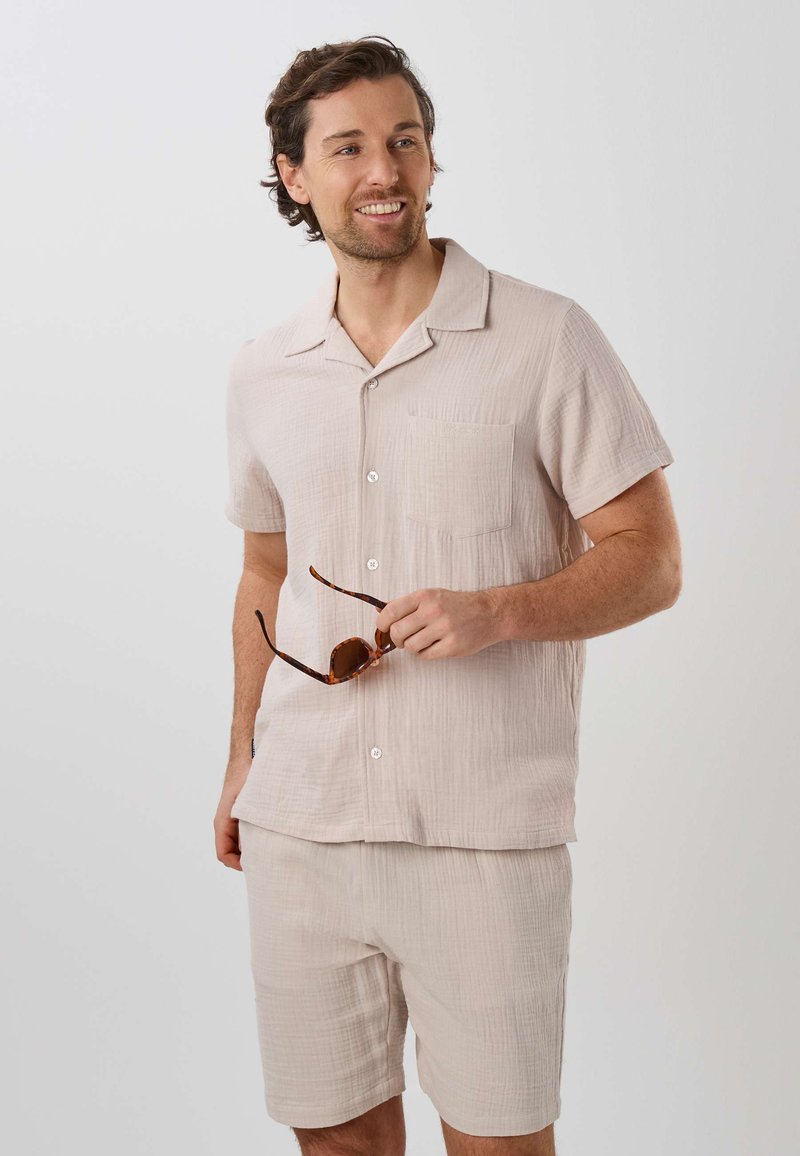 Homme portant une chemise beige clair à manches courtes boutonnée et un short assorti, tenant des lunettes de soleil et souriant en regardant sur le côté.