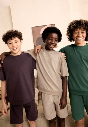 Three boys smiling with arms around each other, wearing matching short-sleeve shirts and shorts in different colors indoors.